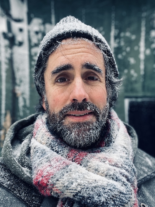 A man smiles at camera while covered in snow frosting his beard, wearing a hat and scarf.