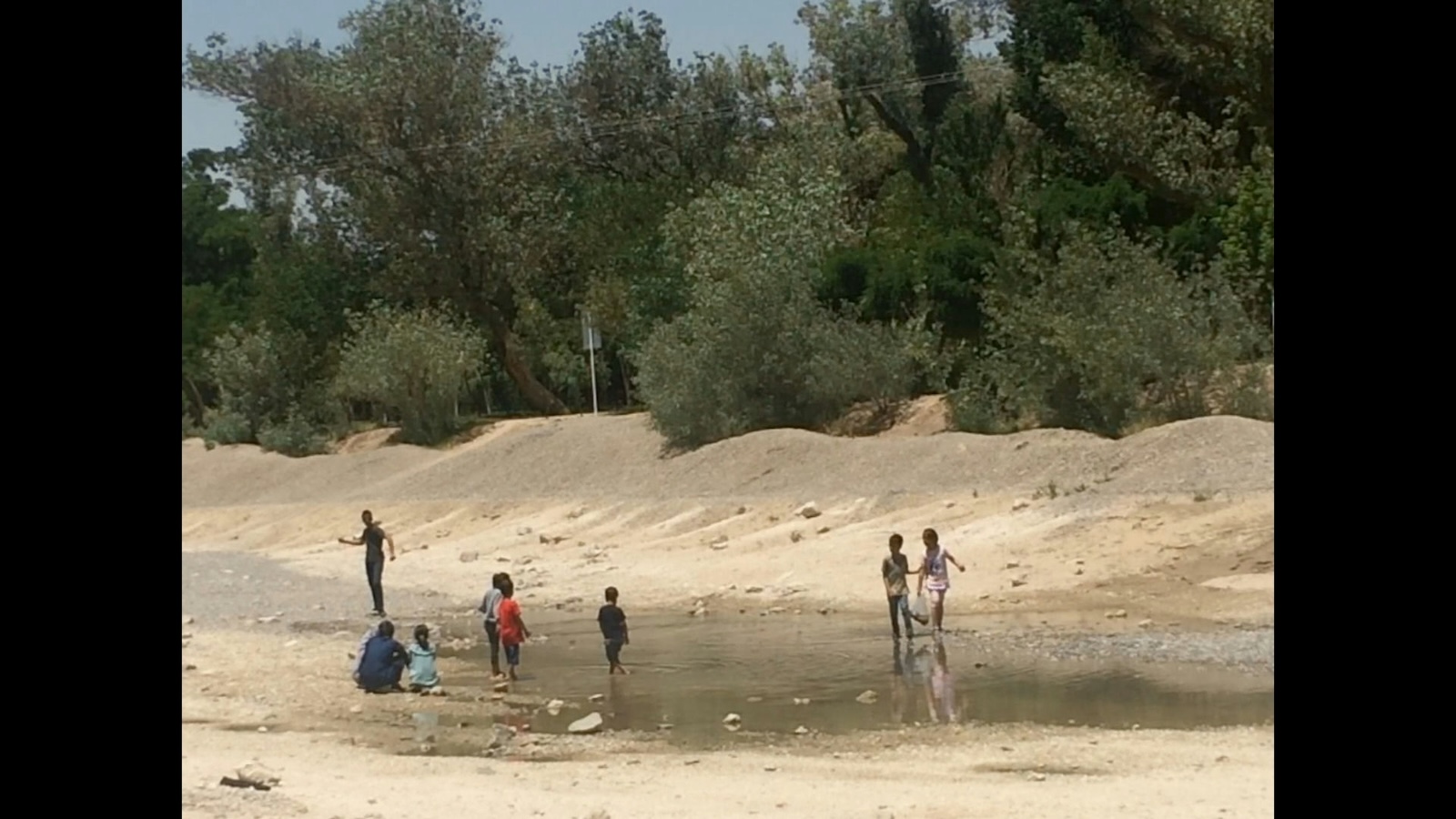 A group of children stand in a shallow reservoir of water in a parched landscape