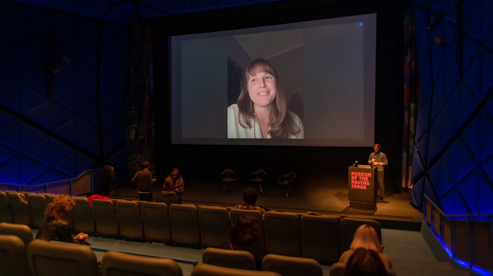 A large movie theater with an image of a woman projected on the screen and a person at a podium on the stage next to the screen