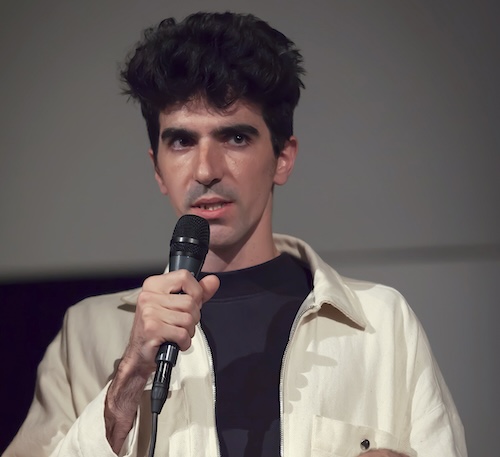 A man in a beige jacket stands talking into a microphone