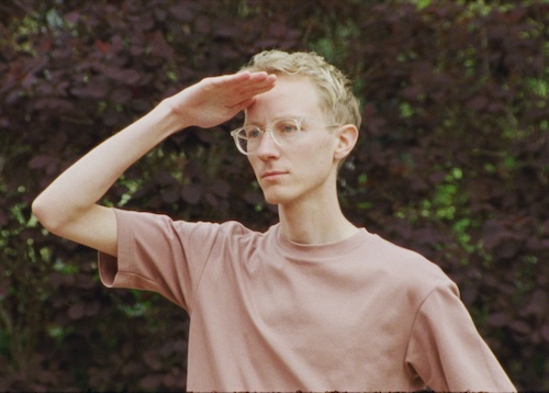 A person with glasses and a pink t-shirt stands outside looking off into distance with a hand covering his forehead to block the sun