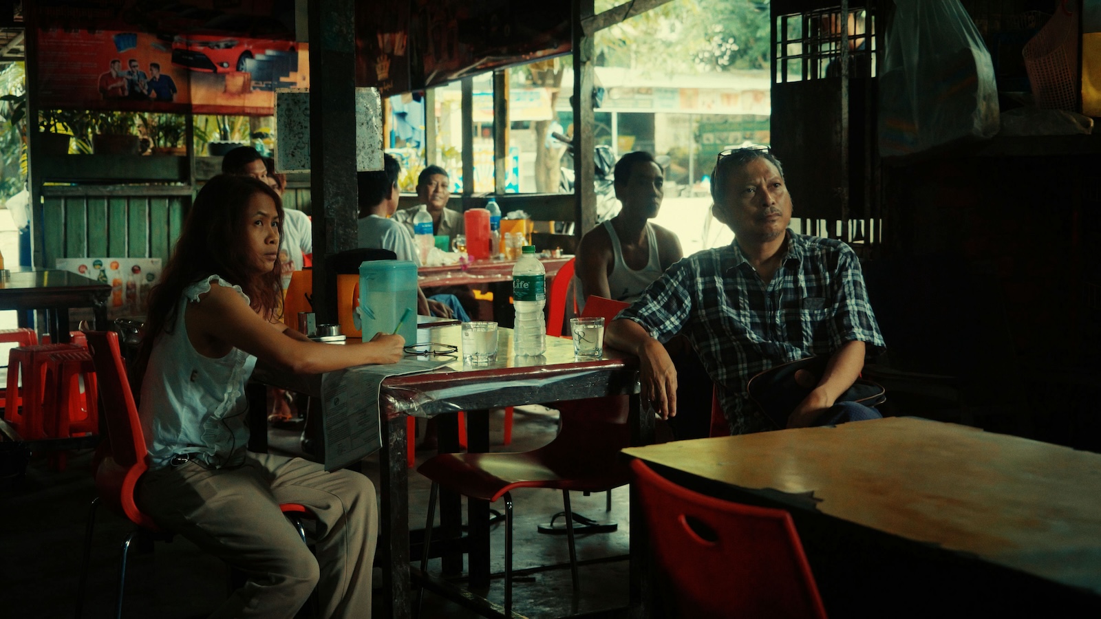 A group of people sit in a cafe in an Asian country looking up at a TV offscreen with serious expressions
