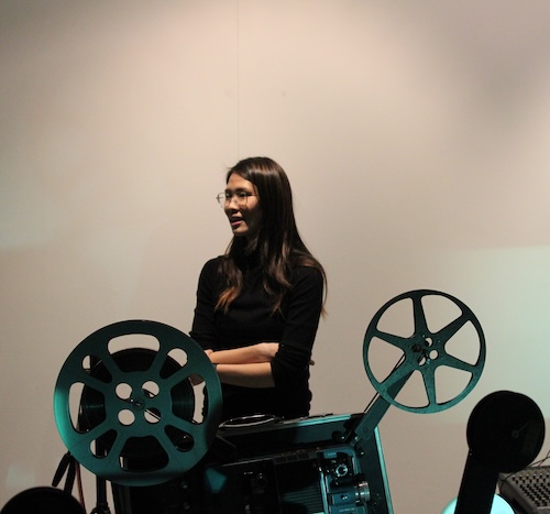 A woman stands with her arms crossed behind a film reel projector in the foreground