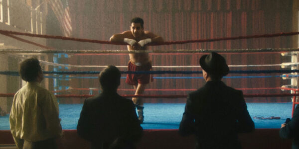 A boxer leans on the ropes in a boxing ring and talks to three men standing on the floor below looking up at him.