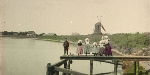 A color-tinted turn of the twentieth century image of people in period clothes standing on a dock over a lake and looking at a windmill