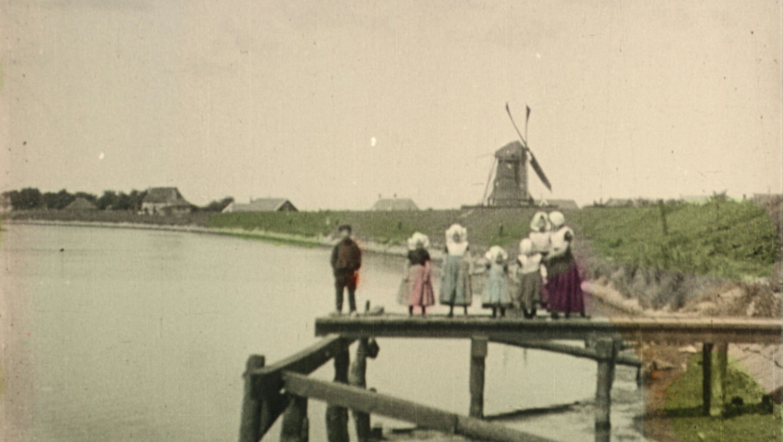 A color-tinted turn of the twentieth century image of people in period clothes standing on a dock over a lake and looking at a windmill