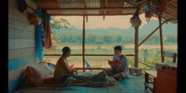 Two men sit in a porch against a bucolic, meditative atmosphere; one reads an open book to the other