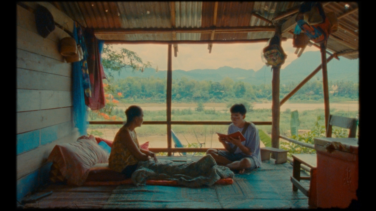 Two men sit in a porch against a bucolic, meditative atmosphere; one reads an open book to the other