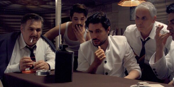 A group of five men wearing white shirts look concerned as they gather around a table and listen to a transistor radio perched on the table in front of them.