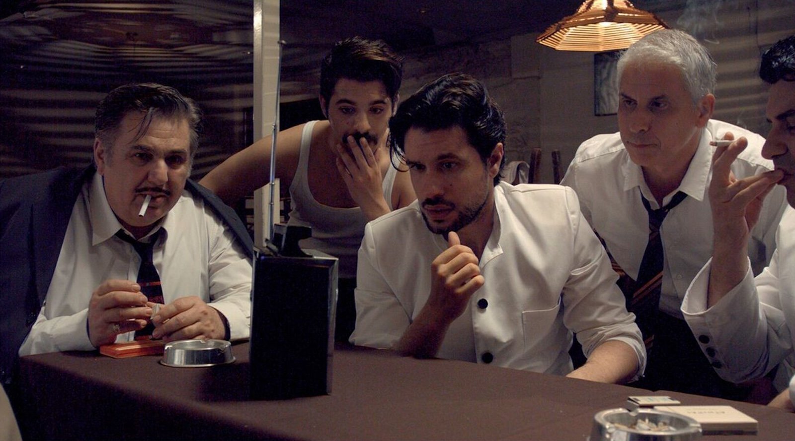 A group of five men wearing white shirts look concerned as they gather around a table and listen to a transistor radio perched on the table in front of them.