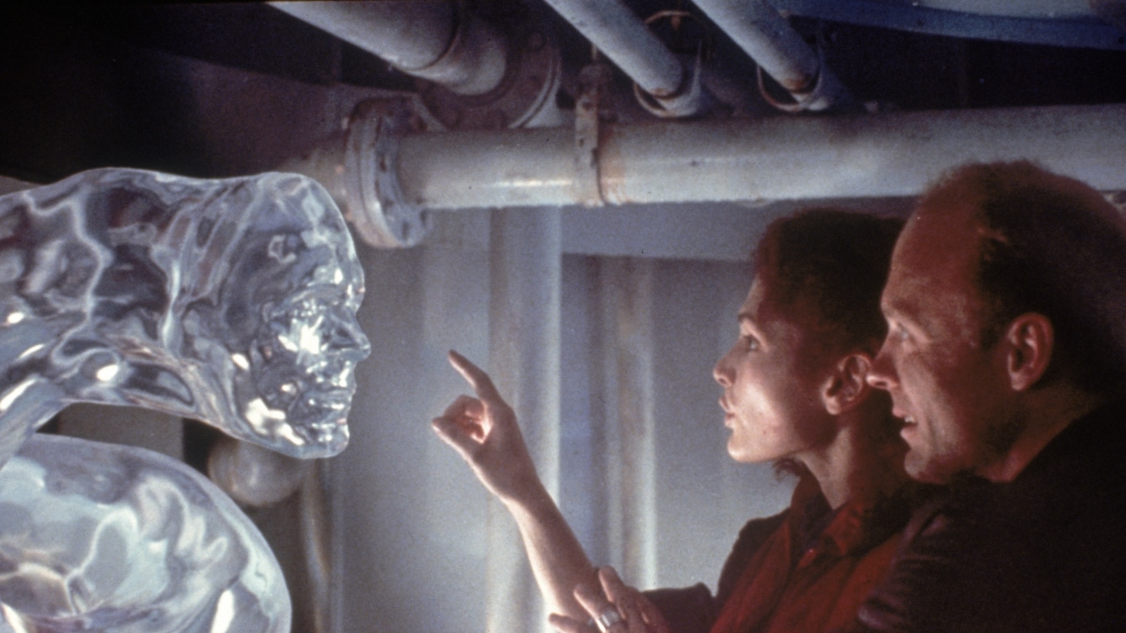 Two people stand in front of a creature with a human face made of water, one pointing her finger at it