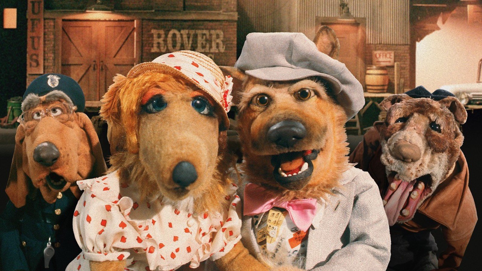 Four puppet dogs dressed in human clothes and hats look at the camera, smiling.