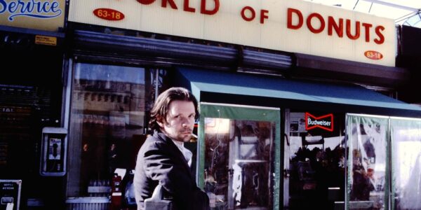 A man with a cigarette in his mouth looks at camera and stands outside a store with the name WORLD OF DONUTS.