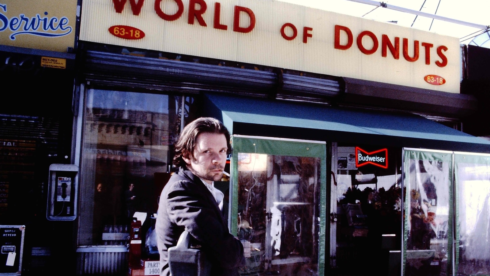 A man with a cigarette in his mouth looks at camera and stands outside a store with the name WORLD OF DONUTS.