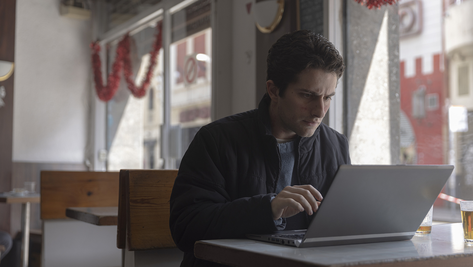A man sits in a restaurant looking concerned at a laptop screen open in front of him.