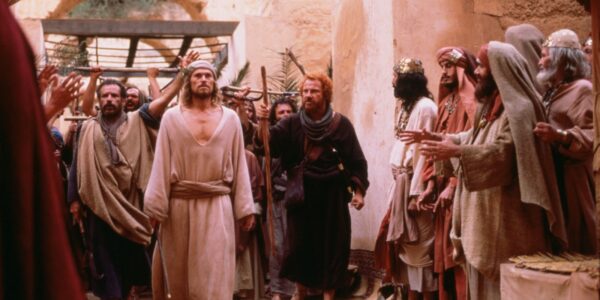 An image from a movie about Jesus Christ, who stands in a loose white robe as he walks in the interior of an ancient city surrounded by other people in ancient Roman peasant dress