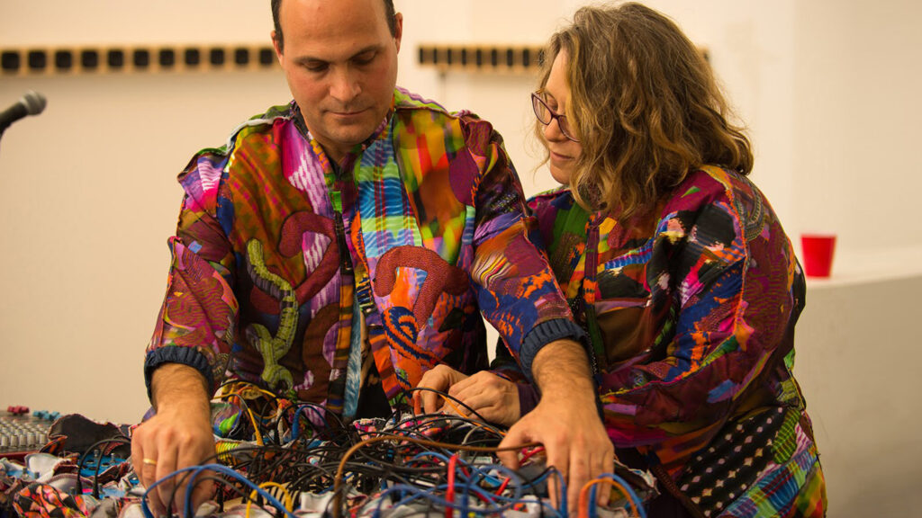 A man and woman in multicolored shirts stand over a control board of multicolored wires