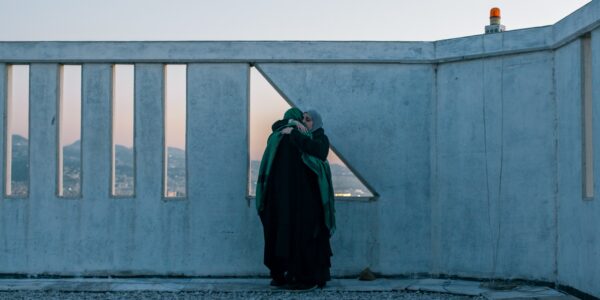 Two women wearing hajibs hug each other while standing on a rooftop, in front of a triangular window, through which we can see a vast mountainous landscape at dusk.