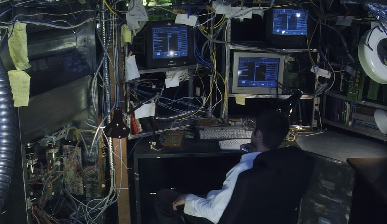 A man, seen from behind, in white shirt its behind a desk surrounded by shelves of messy TV monitors and exposed wiring