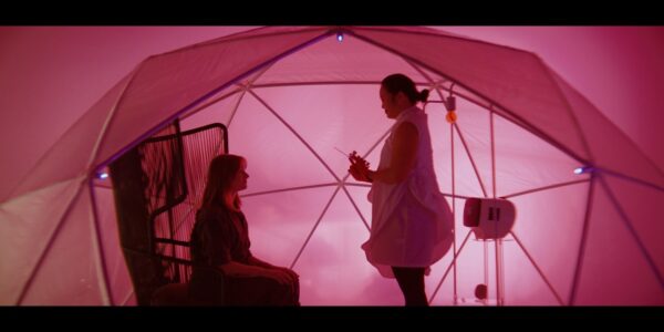 A woman holding a large needle stands over another woman, both under a large pink tent