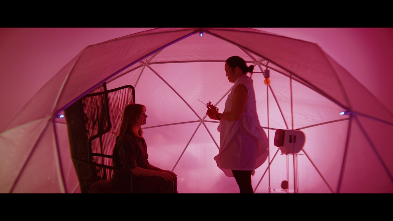 A woman holding a large needle stands over another woman, both under a large pink tent
