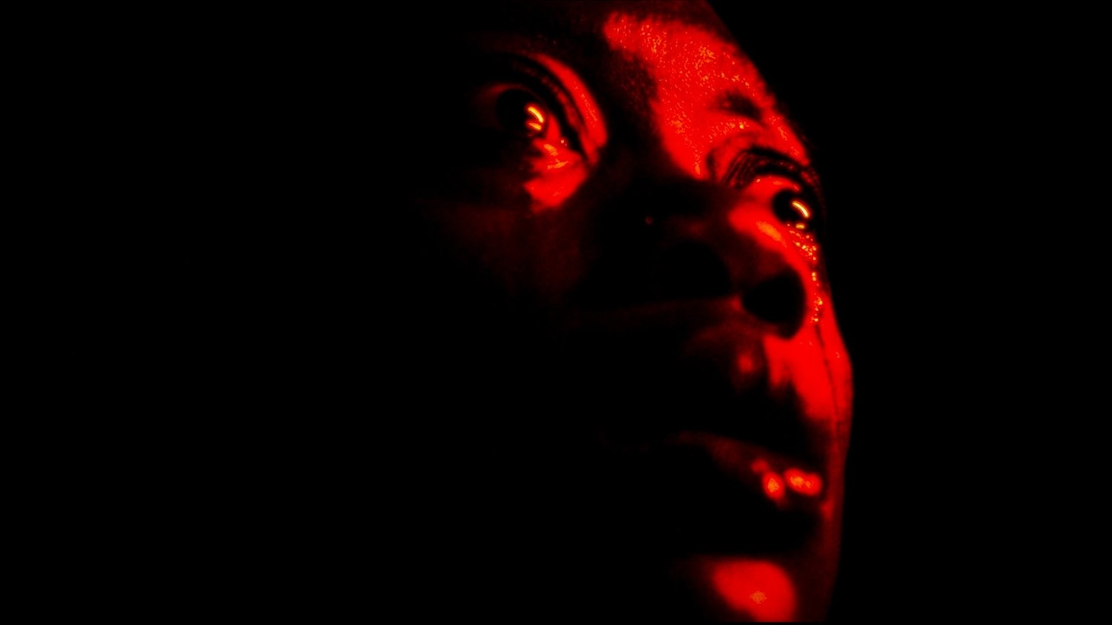 An intense close-up of a face drenched in red looking up and off-screen in panic
