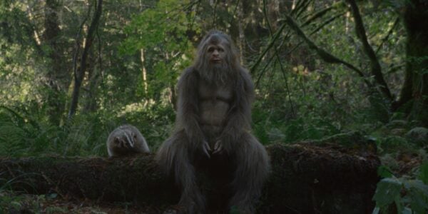 A hairy sasquatch sits on a log in a forest and stares pensively past camera as a badger perches on the log next to it.