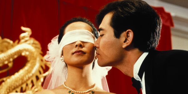 A man ina tuxedo leans over to kiss a blindfolded bride from the side