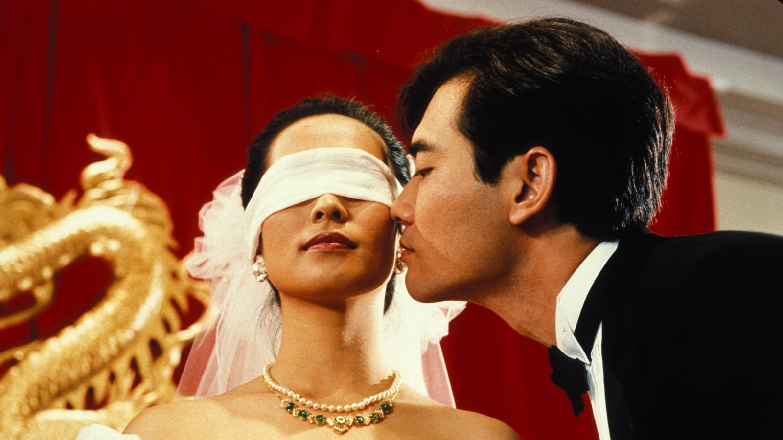 A man ina tuxedo leans over to kiss a blindfolded bride from the side