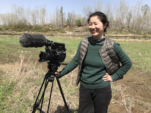 A woman in a vest and green shirt stands in a field holding a video camera on a tripod