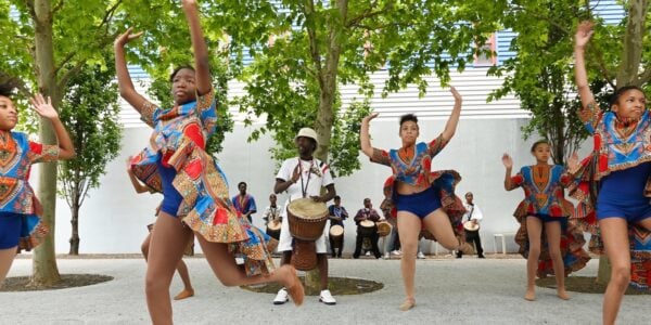 In a courtyard lined with trees, four brown-skinned teenage girls in colorful blue shorts and African-colored shirts dance with their arms in the air as a dark-skinned man in white beats on a drum in the middle of them.
