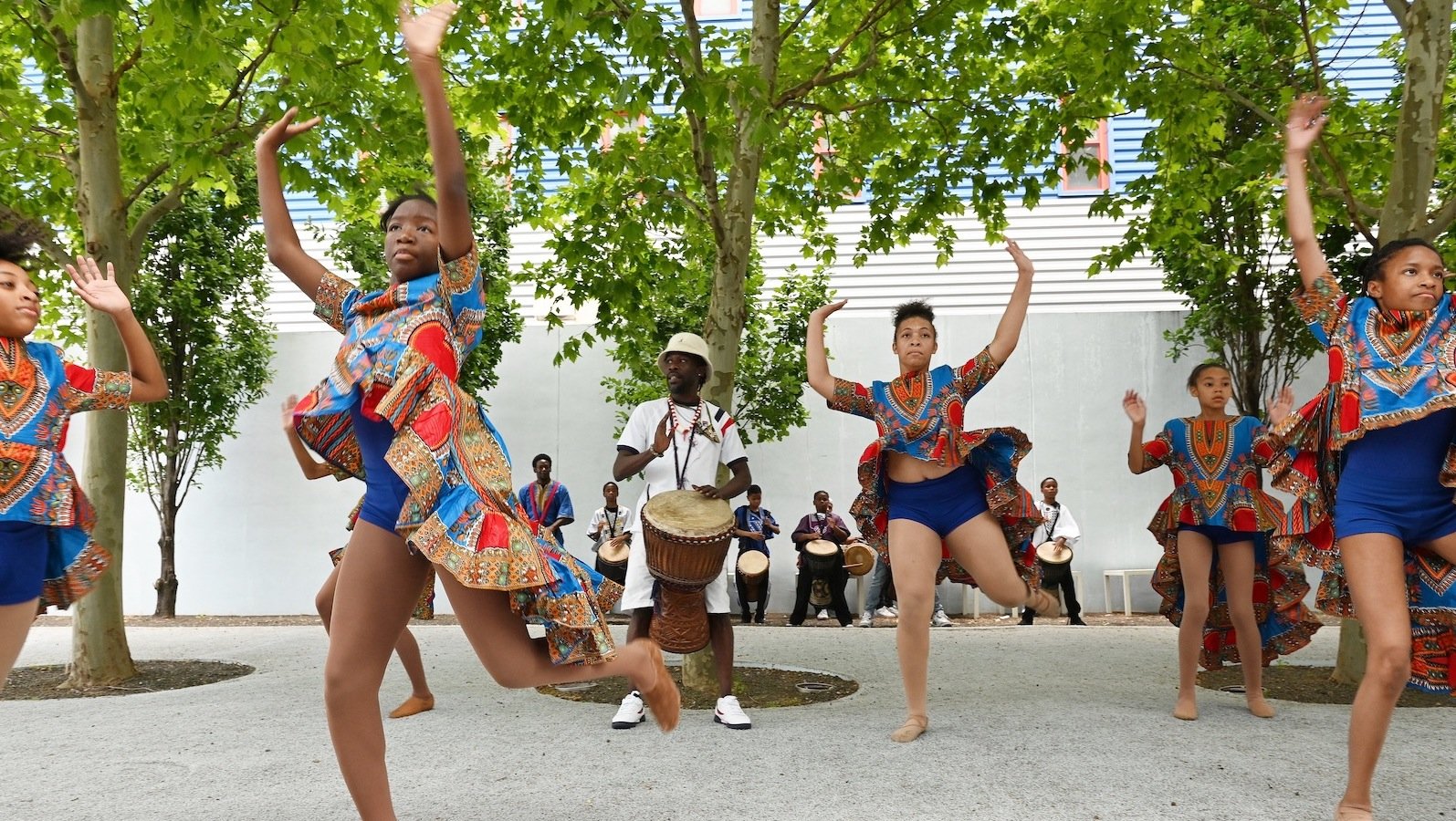 In a courtyard lined with trees, four brown-skinned teenage girls in colorful blue shorts and African-colored shirts dance with their arms in the air as a dark-skinned man in white beats on a drum in the middle of them.