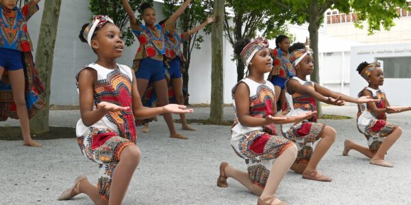 In a courtyard lined with trees, four brown-skinned young girls in white, red and orange, African-style dresses crouch on bended knees with their hands out in performance