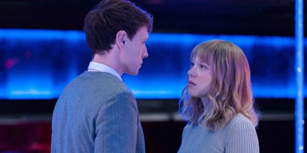 A long-blonde-haired woman in a gray sweater looks up at a man with short dark hair in a gray suit in a modern-looking room with blue neon lights