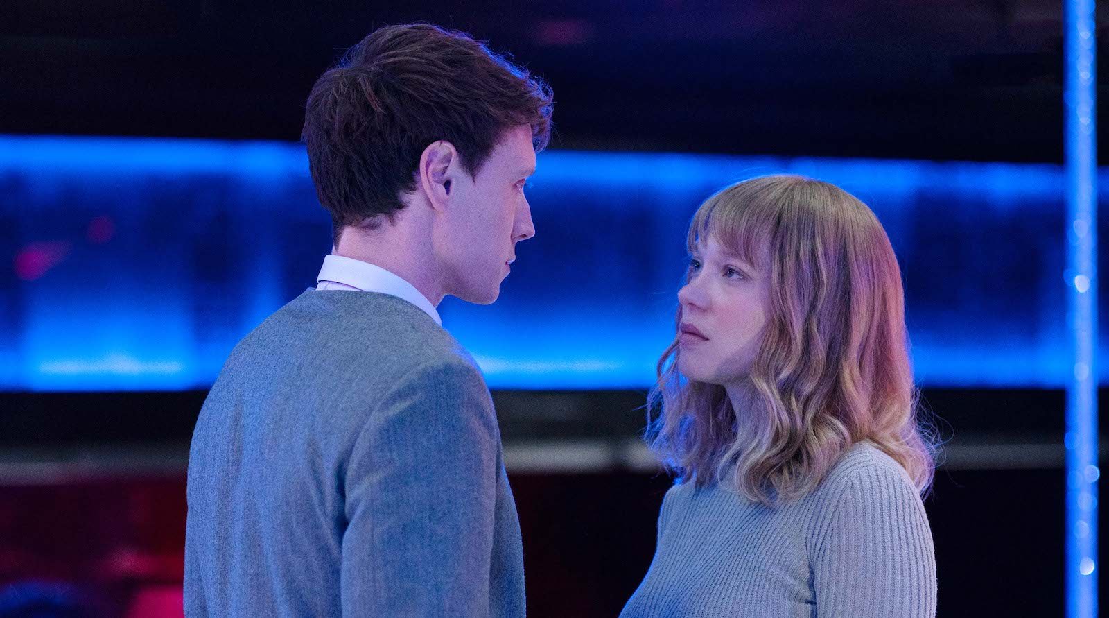 A long-blonde-haired woman in a gray sweater looks up at a man with short dark hair in a gray suit in a modern-looking room with blue neon lights