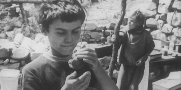 a black and white image of a young boy standing outside cupping an object in his hands, while another boy behind him clutches a wooden post