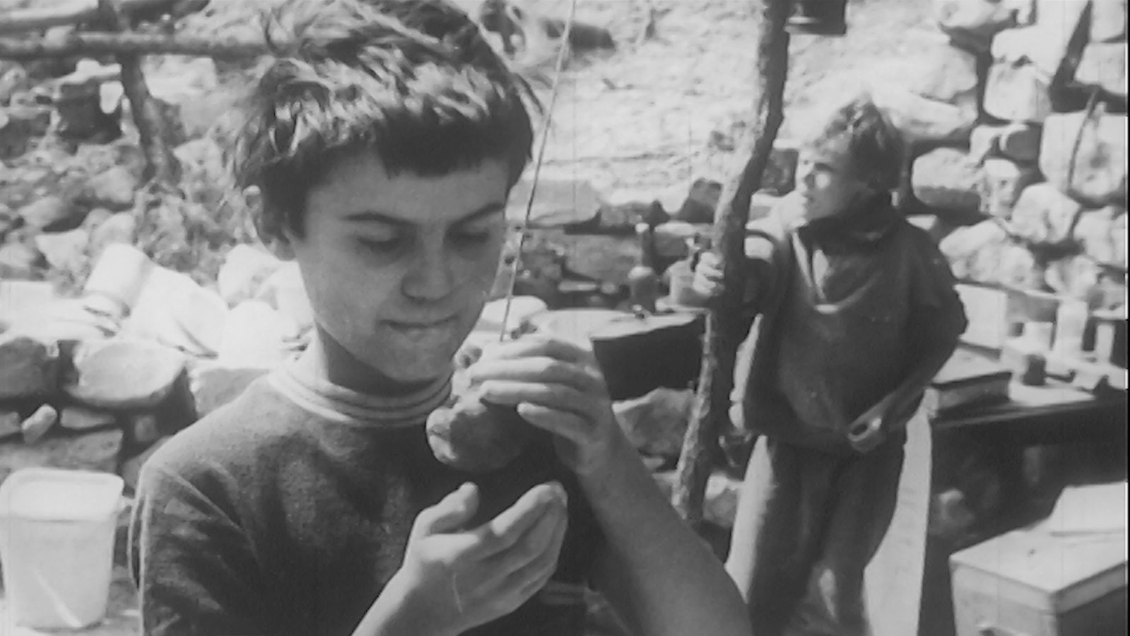 a black and white image of a young boy standing outside cupping an object in his hands, while another boy behind him clutches a wooden post
