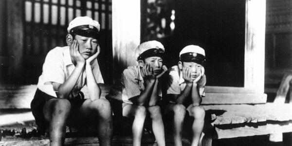 A black and white photo of three Japanese boys in white caps sitting forlornly with their chins in their hands on the edge of a house's front porch.