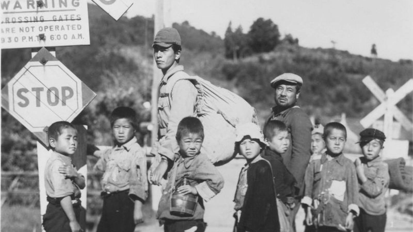 A black and white image of a group of Japanese children and two adults standing in front of a STOP sign at the side of the road looking at the camera forlornly