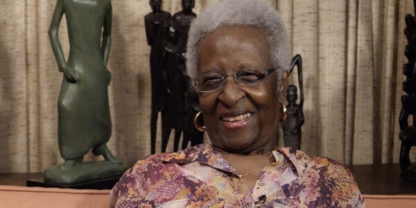 An elderly woman with dark brown skin, white curly hair, gold hoop earrings, and a purple and orange colored shirt sits and smiles, talking to camera, in front of green and black African-style sculptures