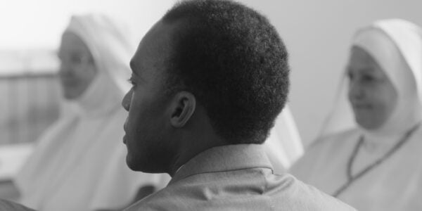 A black and white image of a dark-skinned man, the back of his head facing the camera, with nuns in white habits in the background out of focus