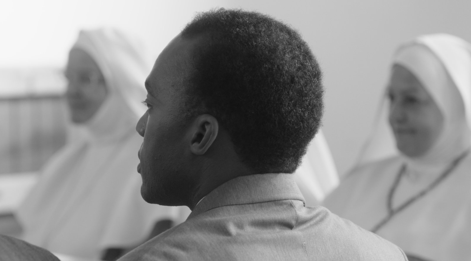 A black and white image of a dark-skinned man, the back of his head facing the camera, with nuns in white habits in the background out of focus