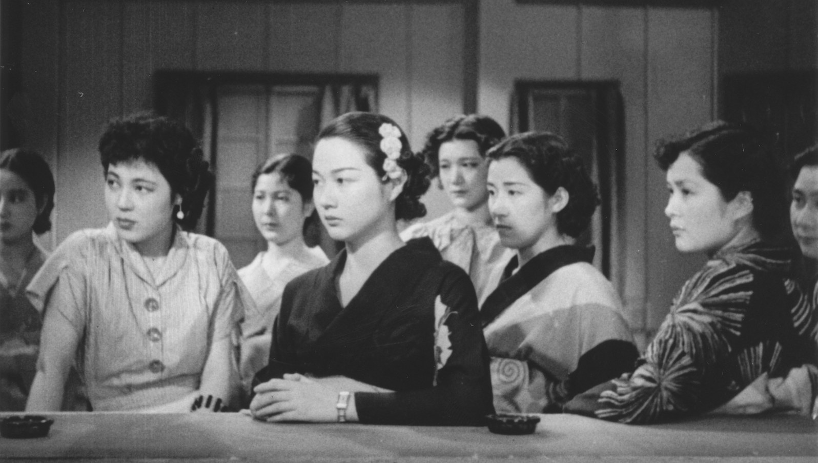 A black and white image of a group of six Japanese women looking left off-screen, the womanin front leaning her elbow on a table, all look serious