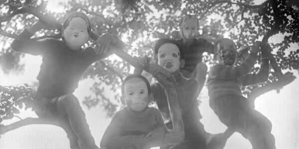 A black and white photo of children in a tree wearing masks and looking down at the camera.