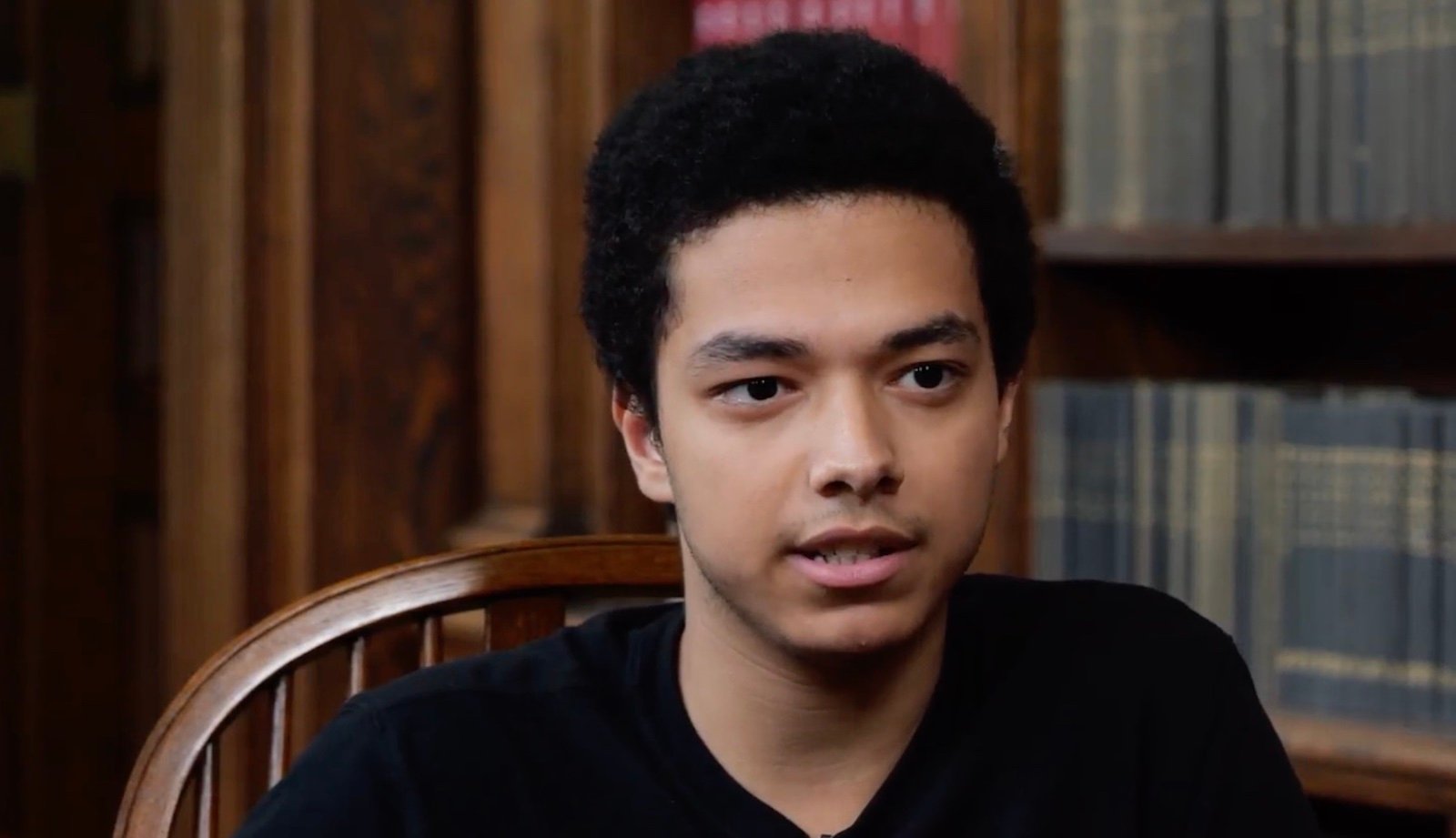 A teenage boy with medium brown skin and dark curly hair sits in a chair talking to camera in front of a bookcase