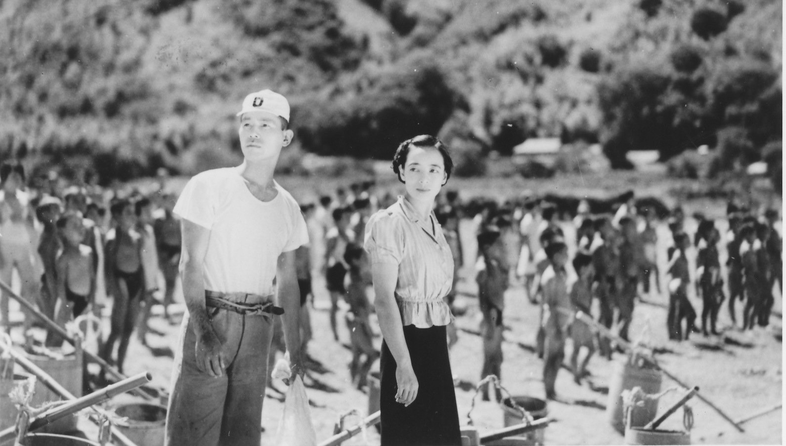 A black and white photo of a Japanese man and woman standing on the shore of a beach, looking off, as lines of children in formation in the background of the shot face the water
