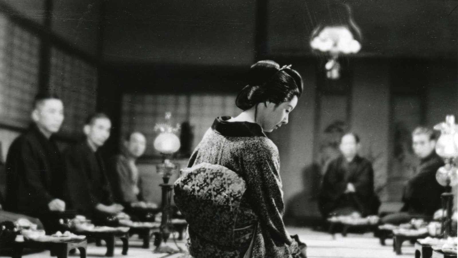 A black and white photo of a woman in traditional Japanese clothes, seen from the back, leaning over a table setting on the floor, which human figures out of focus in the background.