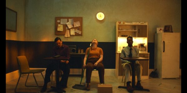 Three people sit in a grim, sickly-colored room in three separate chairs facing thwe camera, with a glowing clock that maybe reads 4:00 on the wall behind them; two men are on either side of a woman, who sits in then middle with her head back in exasperation and holds a pushbroom.