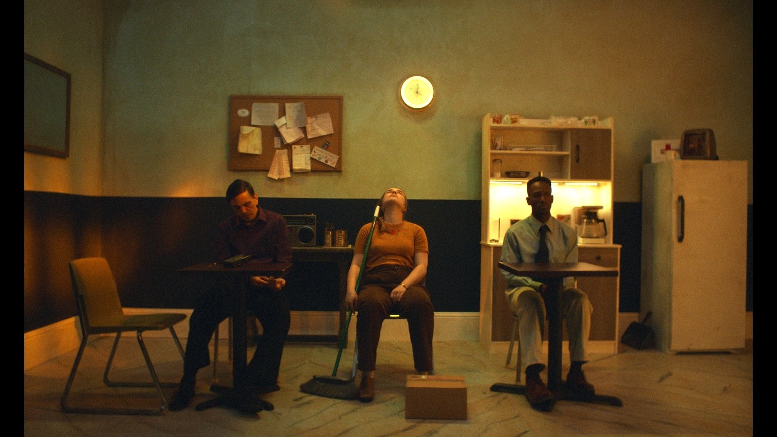 Three people sit in a grim, sickly-colored room in three separate chairs facing thwe camera, with a glowing clock that maybe reads 4:00 on the wall behind them; two men are on either side of a woman, who sits in then middle with her head back in exasperation and holds a pushbroom.