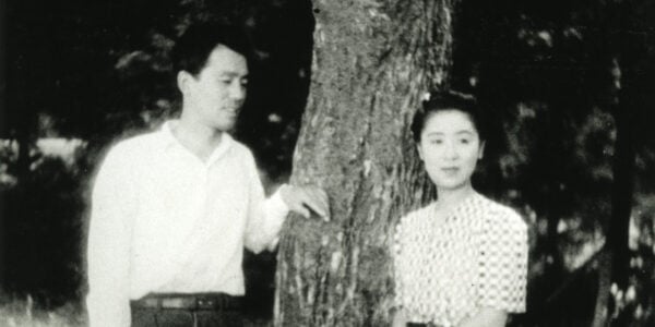 A black and white photo of a Japanese man and woman standing by a tree outside; he looks at her, she looks off past the camera.